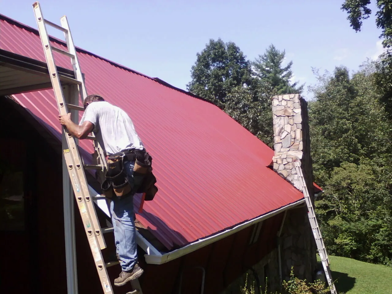 Roofer on a ladder beside a red metal roof and stone chimney — Soffit & Fascia Repair by Roofing Brothers in Sioux Falls